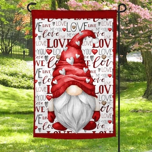 Puede incluir: Una bandera de jardín roja y blanca con un gnomo que lleva un gorro rojo con corazones blancos. La bandera tiene la palabra "love" repetida en rojo y blanco sobre un fondo blanco.