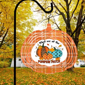 May include: A metal wind spinner with a plaid design in shades of orange and white. The spinner features a circular cutout with a white background and the text "Meet me at the Pumpkin Patch". The cutout also features a graphic of three pumpkins with autumn leaves.