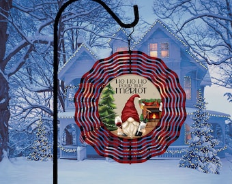 Op de afbeelding: Een rode en zwarte geruite windmolen met een kabouter die een glas wijn en een fles wijn vasthoudt. De kabouter staat voor een open haard en een kerstboom. De tekst op de windmolen is "Ho Ho Ho Pour the Merlot".