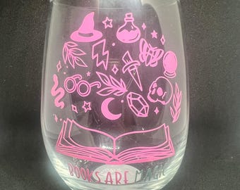 May include: A clear glass wine glass with a pink design of a book, stars, a crescent moon, a skull, a potion bottle, a witch's hat, a sword, a crystal, and glasses. The text "Books are Magic" is printed on the bottom of the glass.