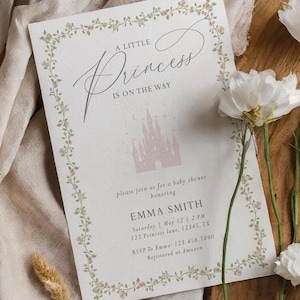 May include: A baby shower invitation with the text "A Little Princess Is On The Way." The invitation features a pink castle graphic and floral border. White flowers and dried plants are arranged around the invitation on a wooden surface.