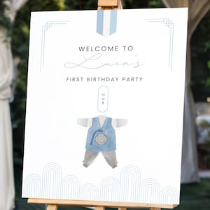 May include: A white welcome sign with blue accents on an easel. The sign reads 'Welcome to [name]'s First Birthday Party'. A blue and white illustration of a baby in traditional Korean clothing is in the center of the sign.