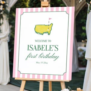 May include: A wooden easel displays a sign with a pink and white striped border. The sign reads "Welcome to Isabelle's first birthday" with a golf course graphic. The event is outdoors with floral decorations.