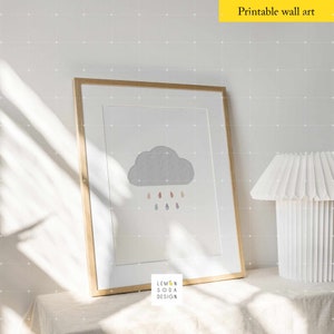 May include: A framed print of a gray cloud with colorful raindrops falling from it. The print is on a white background and has the text "Lemon Soda Design" at the bottom. The print is sitting on a white table with a white lamp next to it.