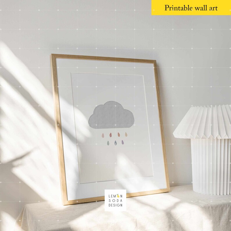 May include: A framed print of a gray cloud with colorful raindrops falling from it. The print is on a white background and has the text "Lemon Soda Design" at the bottom. The print is sitting on a white table with a white lamp next to it.