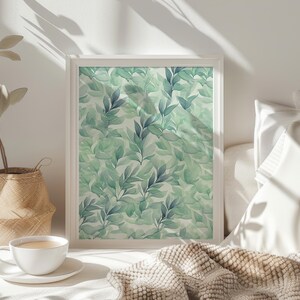 May include: A white framed print with a watercolor design of green and blue leaves on a white background. The print is hanging on a white wall above a bed with white bedding and a brown blanket.