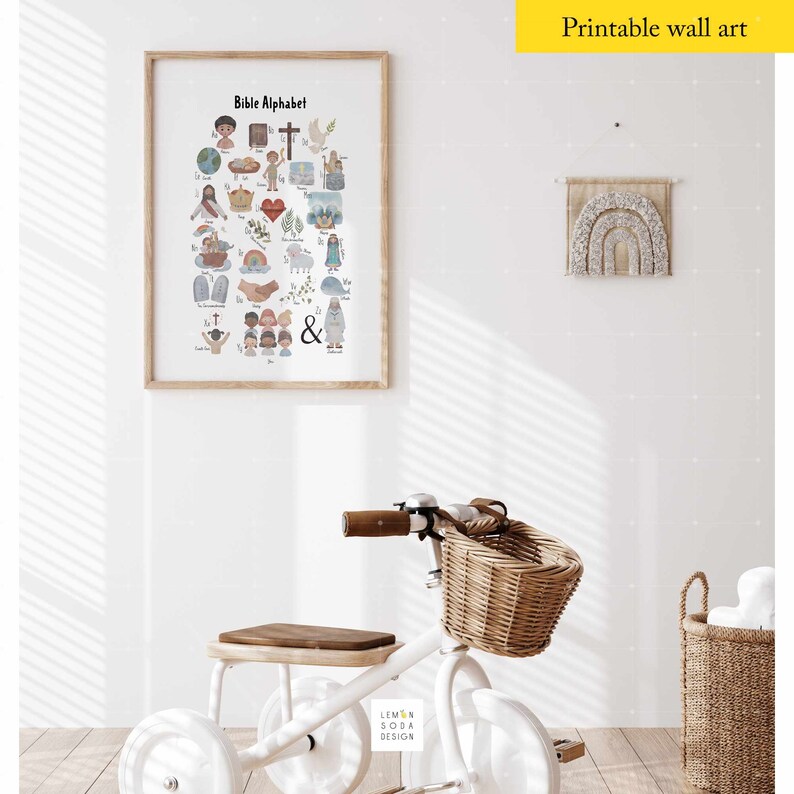 May include: A framed "Bible Alphabet" print with colorful illustrations hangs on a white wall. A white tricycle with a wicker basket and wooden seat sits below the artwork. The text "Printable wall art" is in the top right corner.