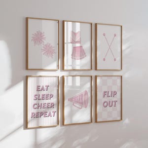 Op de afbeelding: Set van acht roze en witte ingelijste prints met cheerleading-thema's. De prints bevatten pompoenen, een cheerleader-outfit, een megafoon en de woorden "Eat Sleep Cheer Repeat" en "Flip Out".