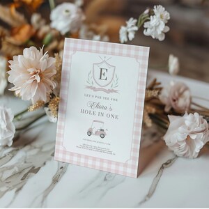 May include: A pink and white checked golf-themed invitation with the text "Let's Par-Tee for Eliora's Hole in One." The invitation features a golf cart illustration and a crest with the letter "E". Decorative flowers surround the invitation.