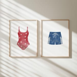 May include: Two framed watercolor prints. One features a red swimsuit with white polka dots. The other depicts blue swim trunks with white accents and a drawstring. Both prints are in light wood frames, perfect for a beach-themed decor.