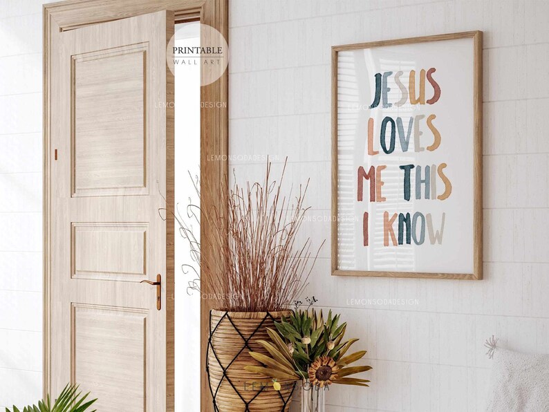 May include: A framed print with the text "Jesus Loves Me This I Know" in colorful, hand-lettered font. The print is in a wooden frame and is hanging on a white wall.