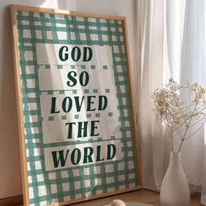 May include: A framed print with a green and white gingham pattern. The print features the text "God so loved the world" in a bold, dark green font.