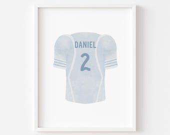 May include: A framed watercolor print of a light blue football jersey. The jersey features the name "DANIEL" above the number "2" in a darker blue. White stripes accent the sleeves and sides of the jersey.