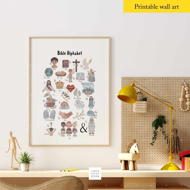 May include: A framed print titled "Bible Alphabet" featuring colorful illustrations for each letter. The artwork includes images of people, objects, and symbols associated with biblical stories and themes. The print is displayed on a desk with a yellow lamp and decorative items.