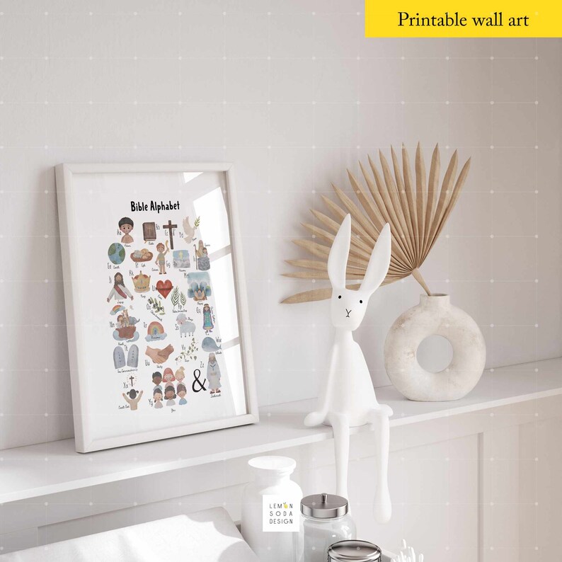 May include: A framed print titled "Bible Alphabet" with colorful illustrations of biblical themes. A white ceramic rabbit figurine and a decorative vase with dried foliage are on a shelf. The text "Printable wall art" is in a yellow rectangle.