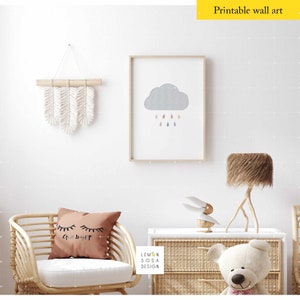 May include: A nursery room interior design with a white wall, a wicker chair with a brown pillow that says "Goodnight" in black text, a framed print of a gray cloud with colorful raindrops, a wooden table with a white ceramic bunny figurine, a teddy bear, and a brown lamp with a fluffy lampshade.