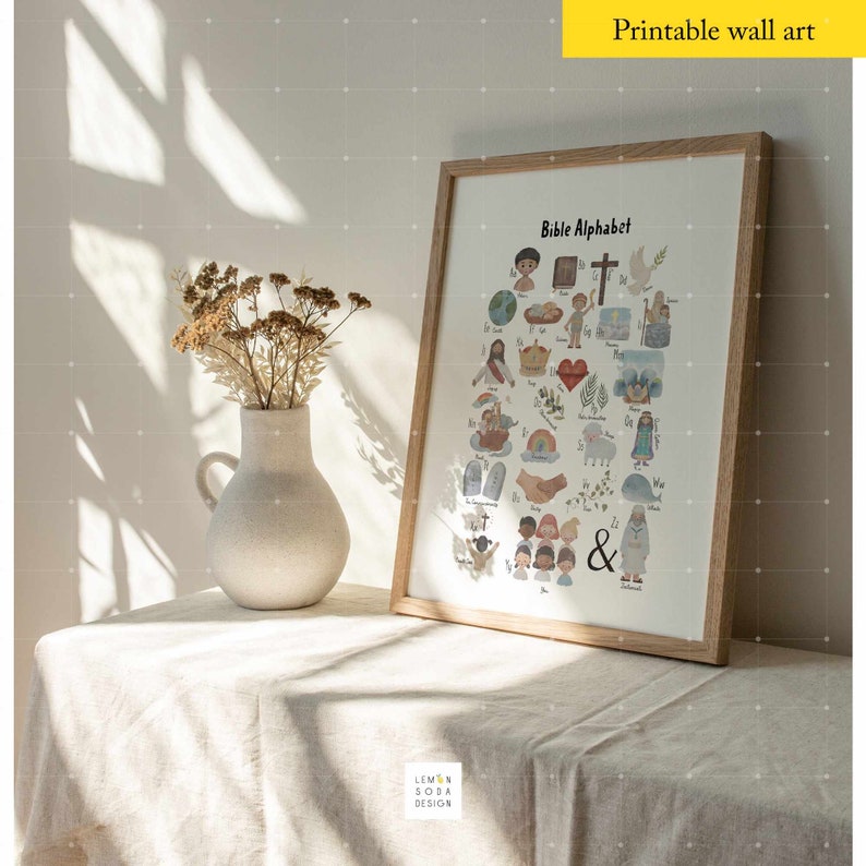 May include: A framed print titled "Bible Alphabet" featuring colorful illustrations for each letter. The artwork is in a light wood frame, displayed on a table with a vase of dried flowers. The text "Printable wall art" is at the top.