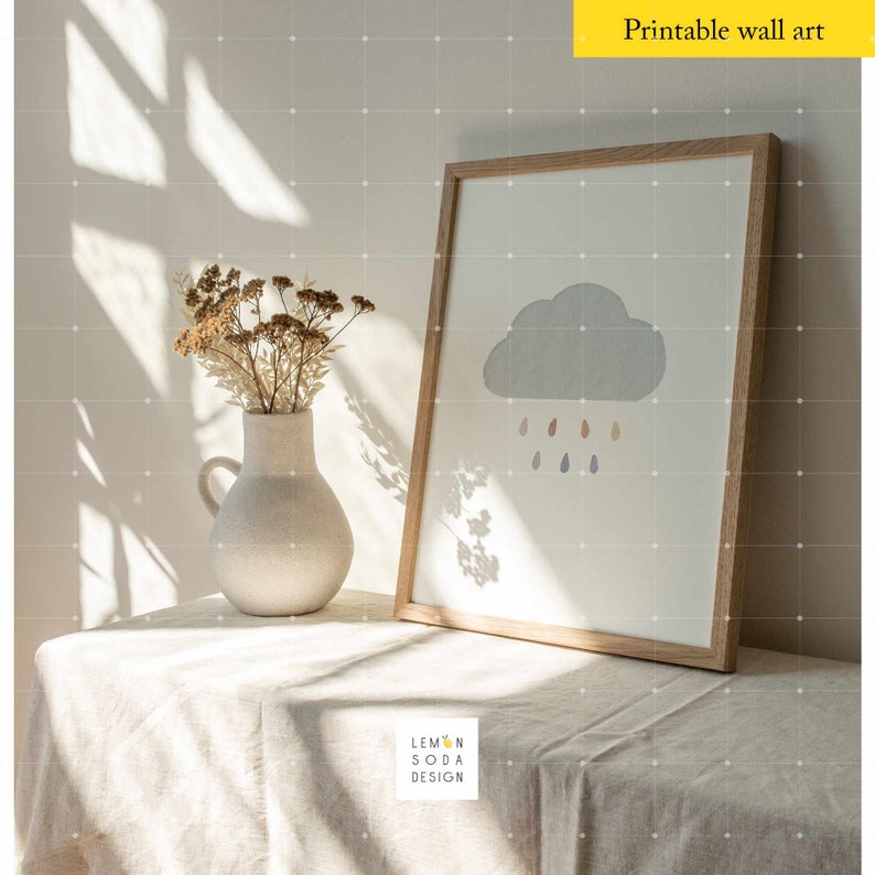 May include: A framed print of a gray cloud with colorful raindrops falling from it. The print is in a light wood frame and is sitting on a white linen tablecloth. The print is titled "Printable wall art".