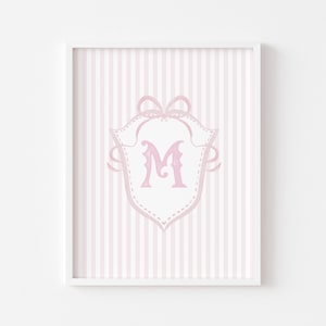 May include: A framed print featuring a pink and white striped background. A pink shield-shaped emblem with a decorative bow and the letter "M" in a script font is centered on the print. The frame is white.