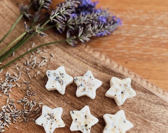 Op de afbeelding: Zes witte stervormige wax melts met lavendel en witte vlokken op een houten oppervlak met lavendeltakken.