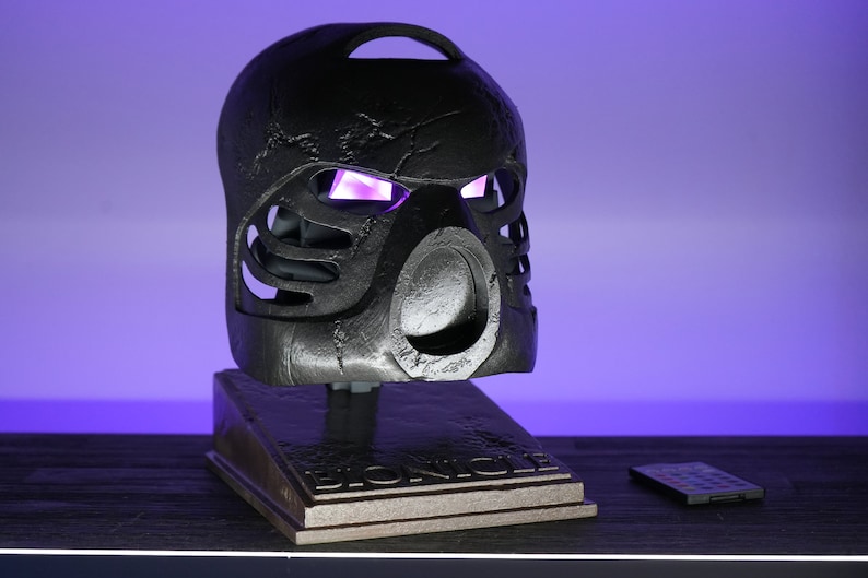 May include: A black Bionicle mask with glowing purple eyes on a stand with the word "Bionicle" on it. The mask is on a dark wooden surface with a remote control on the right side of the image.