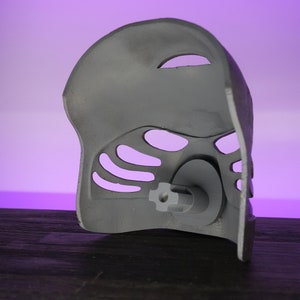 May include: A gray plastic mask with a detailed design and a small hole in the center. The mask has a textured surface and is designed to be worn on the face.