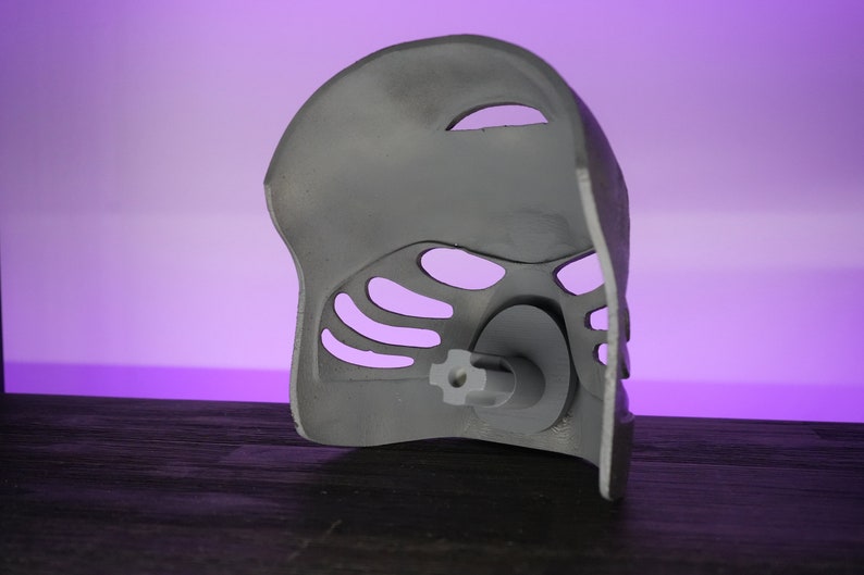 May include: A gray plastic mask with a detailed design and a small hole in the center. The mask has a textured surface and is designed to be worn on the face.