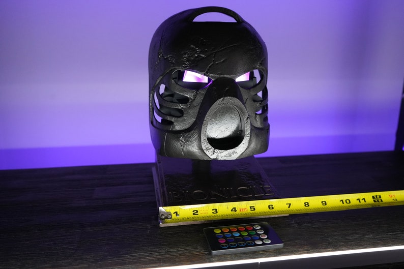 May include: A black Bionicle mask with glowing purple eyes, sitting on a wooden base with the word "Bionicle" printed on it. A tape measure and a remote control with colored buttons are in the foreground.