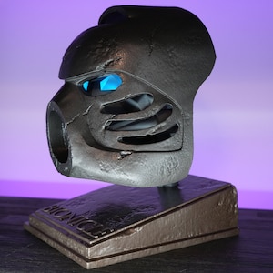 May include: A gray and black Bionicle mask with blue eyes on a gray and brown stand with the word "Bionicle" printed on it.