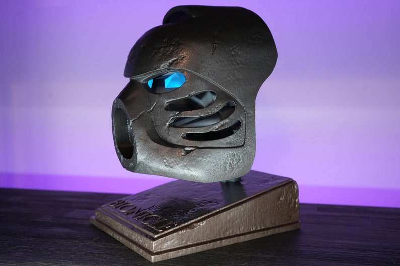 May include: A gray and black Bionicle mask with blue eyes on a gray and brown stand with the word "Bionicle" printed on it.