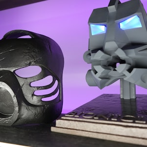 May include: Two black and gray plastic masks, one with glowing blue eyes, on a black and gray base with the word "BIONICLE" printed on it.