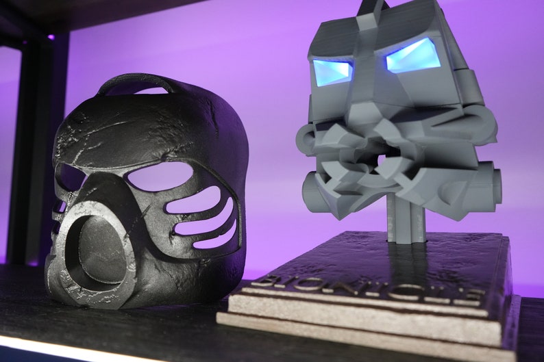 May include: Two black and gray plastic masks, one with glowing blue eyes, on a black and gray base with the word "BIONICLE" printed on it.