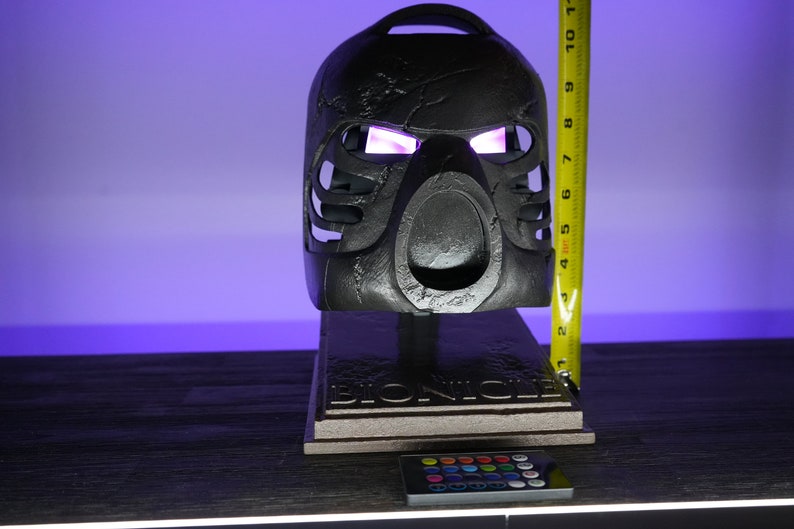 May include: A black Bionicle mask with glowing pink eyes on a stand with a Bionicle logo. The mask is 7 inches tall and comes with a remote control for changing the color of the lights.