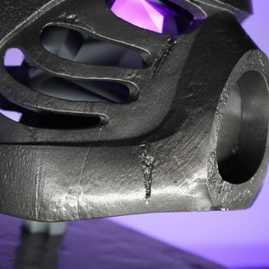 May include: Close-up of a silver-colored 3D printed helmet with a large circular opening on the side. The helmet has a textured surface and appears to be made of plastic.