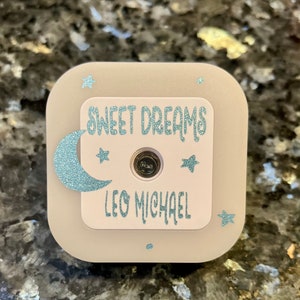 May include: A square, light-colored night light with a central, light pink panel. The panel features the words "SWEET DREAMS" and "LEO MICHAEL" in a glittery, light blue font. A glittery blue crescent moon and stars decorate the night light.