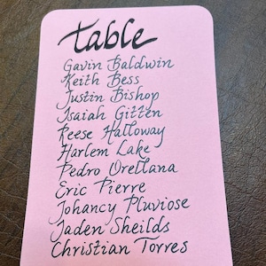 May include: A pink rectangular card with rounded corners, featuring the word "table" in elegant black script. Below, a list of names is handwritten in the same style. The card is set against a dark, textured background.