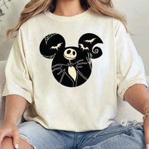 May include: A white t-shirt with a black and white graphic of Jack Skellington from The Nightmare Before Christmas in a Mickey Mouse silhouette. The graphic includes bats and a swirl.