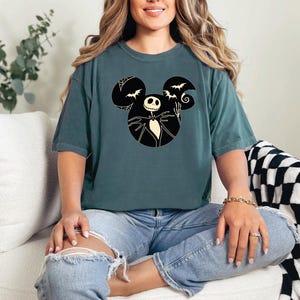 May include: A teal blue t-shirt with a black and white graphic of Jack Skellington from The Nightmare Before Christmas in a Mickey Mouse silhouette. The graphic includes bats and a crescent moon.