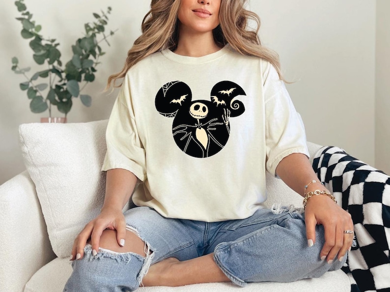 May include: A white t-shirt with a black and white graphic of Jack Skellington from The Nightmare Before Christmas in a Mickey Mouse silhouette. The graphic includes bats and a swirl.