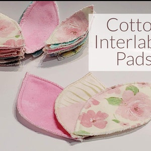 May include: A set of three pink and white cotton interlabial pads with floral patterns. The pads are shaped like leaves and have a white stitched edge. The text "Cotton Interlabial Pads" is written in brown text on a white background.