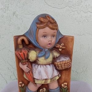 May include: A ceramic figurine of a girl wearing a blue and yellow dress and a blue bonnet. She is holding a red umbrella and a basket. The figurine is standing on a brown base with green grass and yellow flowers.