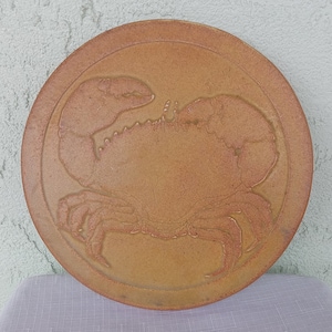 May include: A round, terracotta-colored ceramic plate with a raised design of a crab. The crab is in the center of the plate and is facing to the right. The plate has a slightly raised rim.