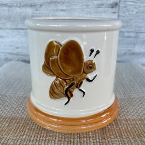 May include: A ceramic, cylindrical container with a bee design. The container is primarily off-white with a brown and orange base and a brown bee with black details. The bee is in relief, adding texture to the design.
