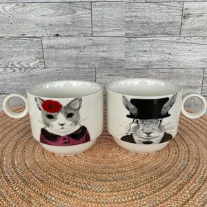 May include: Two white ceramic mugs with circular handles. The left mug features a gray cat wearing a red flower and a dark pink garment. The right mug features a gray rabbit wearing a black top hat and a monocle over one eye, with a dark suit.