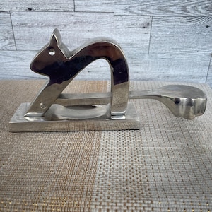 May include: A silver-colored nutcracker in the shape of a squirrel. The nutcracker has a rectangular base with a lever and a squirrel-shaped head. The lever extends to a round, flat surface.