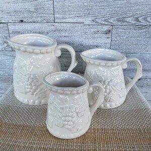 May include: Three white ceramic pitchers with embossed grape and leaf designs. The pitchers vary in size, with handles and decorative rims. They are arranged on a woven beige placemat against a light gray wood background.