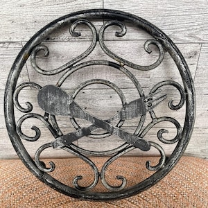 May include: A round, black metal wall decor with a decorative design of intertwined swirls. A fork and spoon are crossed in the center of the design.