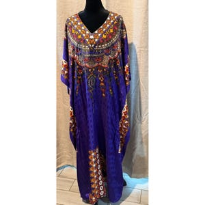 May include: A floor-length purple kaftan featuring a V-neckline and a vibrant, multi-colored geometric and floral pattern around the collar and down the sides. The fabric has a subtle, repeating square texture throughout the garment's surface.