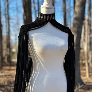 Op de afbeelding: Een zwart macramé schouderstuk met een hoge kraag en zilverkleurige metalen accenten. Het kledingstuk is gemaakt van lange, verticale zwarte koordfranjes die vanaf de halslijn naar beneden over de romp vallen, wat een gelaagd effect creëert.