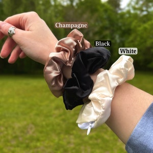 May include: Three satin scrunchies in champagne, black, and white. The scrunchies are displayed on a wrist. The champagne scrunchie is a light, warm beige color. The black scrunchie is a deep, solid black. The white scrunchie is a soft, creamy white.